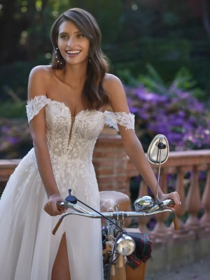 Robe de mariée coupe droite au style romantique, réalisée en tulle, dentelle et strass. Il présente un décolleté profond, des bretelles amovibles et une jupe fendue sur le côté. Une robe de mariée Adriana Alier